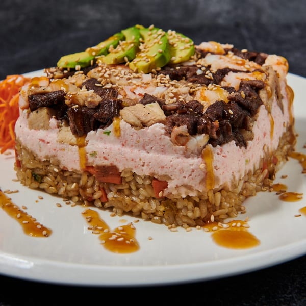 Sushi cake with rice, pink filling, meat, shrimp, avocado, and sesame seeds on a white plate.