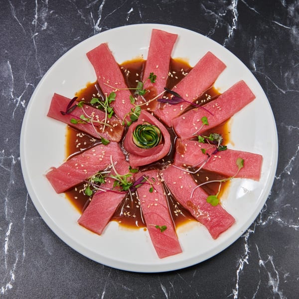 Tuna sashimi slices arranged in a circle with a cucumber rose, sesame seeds, and microgreens.