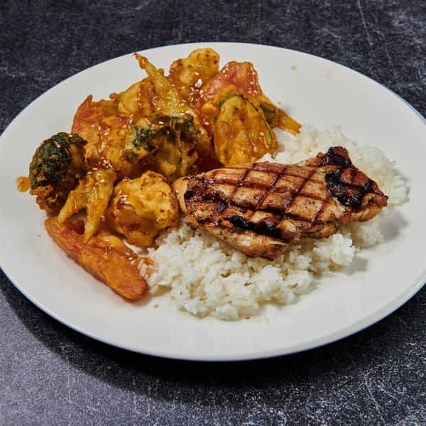 Grilled chicken, tempura vegetables, and white rice on a white plate, drizzled with sauce and sesame seeds.