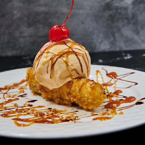 Fried banana dessert topped with vanilla ice cream, caramel, chocolate drizzle, and a cherry.