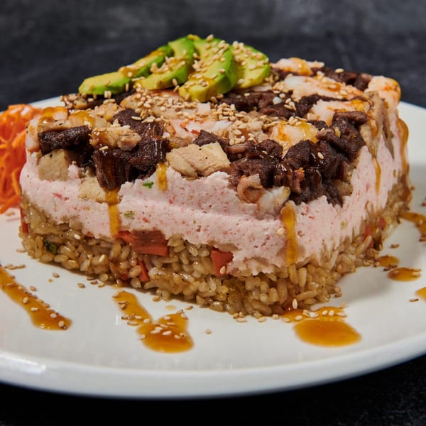 Sushi cake with rice, pink crab salad, beef, shrimp, avocado, sesame seeds, and sauce.