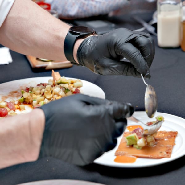 Chef in black gloves plating a sushi dish with diced ingredients on a crispy base.