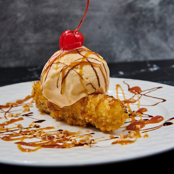 Fried banana dessert topped with vanilla ice cream, caramel, chocolate drizzle, and a cherry.