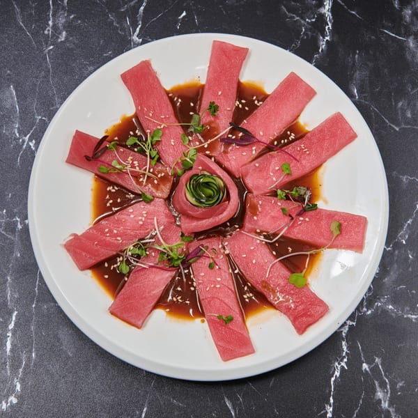 Tuna sashimi slices arranged in a circle with a cucumber rose, sesame seeds, and microgreens.