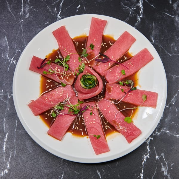 Tuna sashimi slices arranged in a circle with a cucumber rose, sesame seeds, and microgreens.