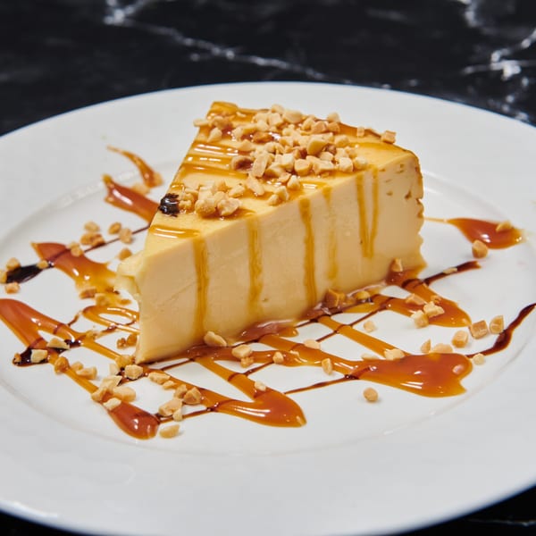 A slice of flan topped with caramel drizzle and chopped peanuts on a white plate.
