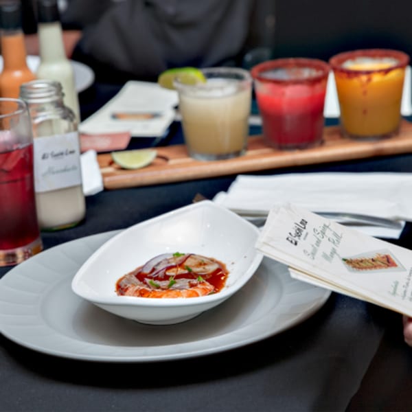 A white bowl of sushi with red sauce and green garnish, alongside drinks and menus.