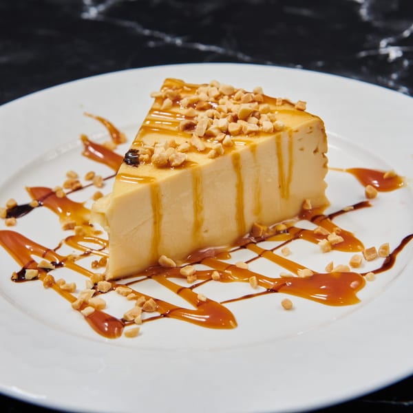 Slice of flan dessert with caramel drizzle and chopped peanuts on a white plate.