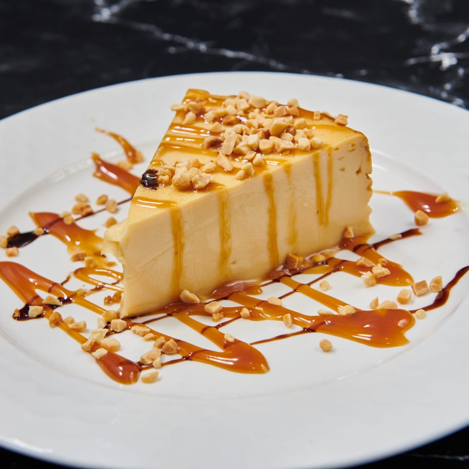 Slice of flan dessert with caramel drizzle and chopped peanuts on a white plate.