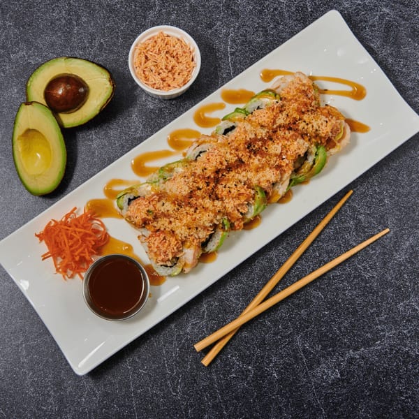 Sushi rolls with avocado, sauce, and crunchy topping, with avocado halves and chopsticks.